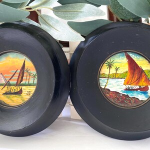 May include: Two round, black framed miniature paintings. One depicts a sailboat on a body of water with a red sail, palm trees, and a sunset. The other shows a sailboat with pyramids in the background.