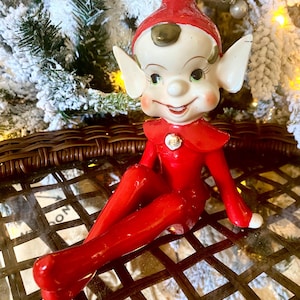 May include: A vintage Christmas elf figurine. The elf is dressed in a bright red outfit and hat, with a white face, large pointed ears, and a smiling expression. The figurine is sitting on a brown wicker surface, with a snowy Christmas tree in the background.