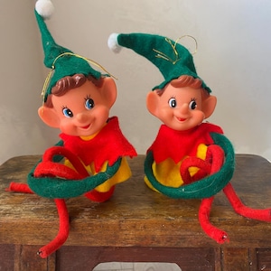 Puede incluir: Dos figuras de duendes navideños vintage. Cada duende tiene un gorro verde con un pompón blanco, un cuello rojo y una camisa amarilla. Los duendes tienen piernas rojas y están sentados sobre una superficie de madera.