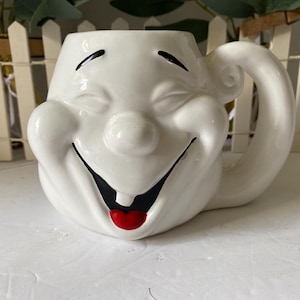 Könnte beinhalten: Eine weiße Keramiktasse in Form eines lächelnden Gesichts. Die Tasse hat schwarze Augen, ein breites schwarzes Lächeln und eine rote Zunge. Der Griff ist gebogen und die Tasse steht auf einer weißen Oberfläche.