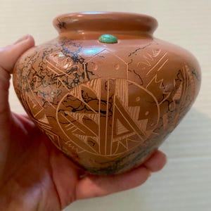 May include: A terracotta-colored ceramic pot featuring intricate etched geometric patterns and black horsehair-style markings. A small, round, polished turquoise stone is embedded near the rim of the vessel, which has a wide, rounded body and a short, narrow neck.