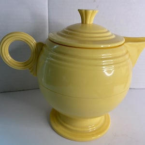 Puede incluir: Una tetera de cerámica de color amarillo pálido con tapa y boquilla. La tetera tiene un cuerpo redondo, una base de pedestal y un asa circular. La tapa está rematada con un pequeño adorno decorativo. La tetera es probablemente vintage o retro.
