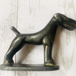 Puede incluir: Una figura de perro de metal gris oscuro. El perro está de pie sobre una pequeña base ovalada. La figura tiene un acabado metálico liso y es un objeto decorativo.
