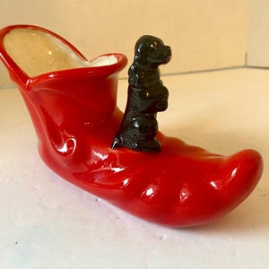 May include: A red ceramic shoe-shaped planter with a black dog figurine. The inside of the shoe is beige. The planter is a decorative item. The shoe's length is approximately 15 cm.