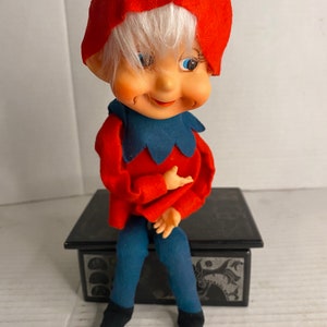 Puede incluir: Una muñeca de elfo de Navidad vintage con un gorro rojo, cabello blanco, ojos azules y un atuendo rojo y azul. El elfo está sentado en una caja negra con un diseño plateado.