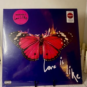 May include: Maroon 5's "Love is Like" album cover. The album art features a vibrant red butterfly with black and white accents against a deep blue background. The album title is written in white script. A sticker indicates this is an exclusive edition.