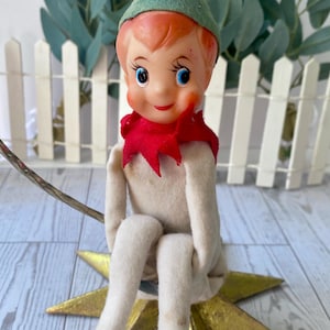 Puede incluir: Una muñeca elfo navideña vintage con un gorro de fieltro verde, un cuello rojo y una cara pintada con ojos azules. El elfo está sentado en un adorno dorado en forma de estrella, una decoración navideña clásica. El fondo incluye una valla blanca y vegetación.