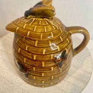 May include: A brown ceramic honey pot shaped like a beehive with a textured basket-weave pattern. The lid features a sculpted bee on top, and the sides of the pot are decorated with two additional raised bee figures. It includes a handle and a spout.
