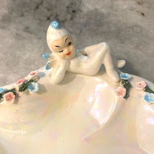 Puede incluir: Figura de cerámica vintage de un hada reclinada sobre un plato nacarado. El hada es blanca con detalles azules y labios rojos. El plato está decorado con flores rosas y azules.