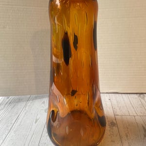 Peut inclure: Un vase en verre ambré de forme élancée, avec une base évasée et un col étroit. La surface est texturée avec des indentations ovales allongées. Des accents brun foncé sont visibles dans le verre. Le vase est vide. Objet décoratif.