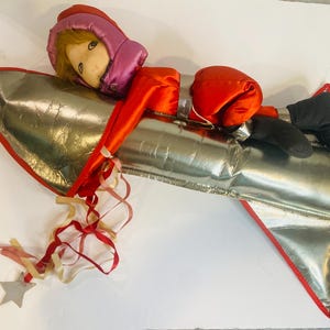 May include: A vintage toy rocket with a doll astronaut. The rocket is silver with red trim, and the astronaut wears a red and pink space suit with black boots. A star-shaped charm hangs from the rocket.