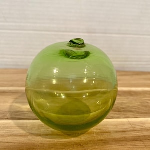 May include: A round, green glass object with a small, dark green top. The glass has a slight yellow tint and sits on a wooden surface. The object is spherical and appears to be a decorative piece or a small container.