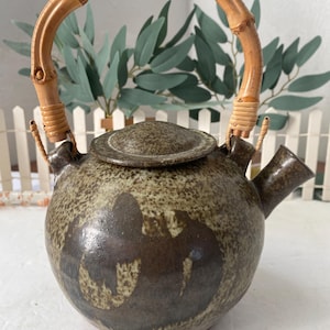 Könnte beinhalten: Eine Keramik-Teekanne mit Bambusgriff. Die Teekanne hat eine gesprenkelte braune und beige Farbe mit einem dunkelbraunen Design auf der Vorderseite. Der Griff ist gebogen und mit geflochtenem Bindfaden an der Teekanne befestigt. Der Deckel ist rund.