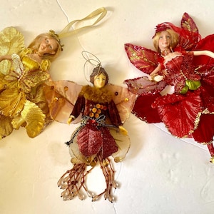 Peut inclure: Trois décorations de fées avec des ailes. Une fée porte une robe dorée avec des ailes dorées, une autre porte une robe rouge avec des ailes rouges et la dernière porte une robe bordeaux avec des ailes brunes.