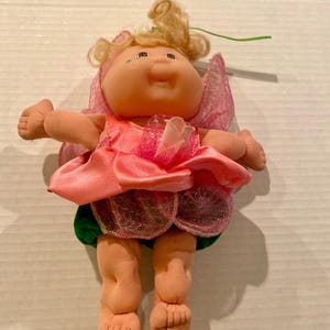 May include: A vintage Cabbage Patch Kids doll dressed as a fairy. The doll has blonde curly hair, a peach-colored face, and is wearing a pink satin dress with matching wings. The doll's skirt is green and the wings are a sheer pink material.