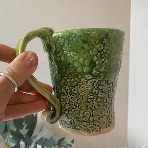 Op de afbeelding: Een handgemaakte groene keramische mok met een bloemenpatroon. De mok heeft een reliëfpatroon van bloemen en bladeren. De handgreep is ook groen en heeft een organische vorm. De mok wordt door een hand vastgehouden.