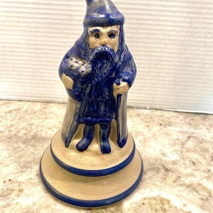 May include: A ceramic wizard figurine in blue robes and hat, holding a staff and a bundle. The wizard has a long white beard and stands on a beige base with blue trim. The figurine is approximately 15 cm tall.