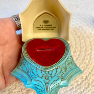 May include: A heart-shaped ring box with a turquoise exterior featuring gold-colored filigree patterns. The interior is lined with red velvet and includes a cream-colored lid with the text V. J. CLEMENS JEWELER & OPTOMETRIST CAPE GIRARDEAU, MO.