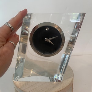 May include: A clear, angular glass desk clock featuring a circular black face with a silver-toned bezel and hands. The clock face includes a single silver dot at the twelve o'clock position and the brand name Movado printed in small silver text.