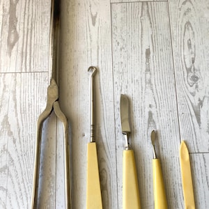 Peut inclure: Ensemble d'outils anciens en métal et ivoire, comprenant un fer à friser en métal, un crochet, deux couteaux et une petite spatule. Les outils sont disposés sur une surface claire imitation bois.