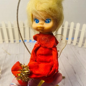 May include: A vintage Christmas angel doll with a blonde hairstyle, blue eyes, and a red velvet dress. The doll has gold tinsel accents on its halo and shoes. The doll is held in a hand, with a star-shaped base.