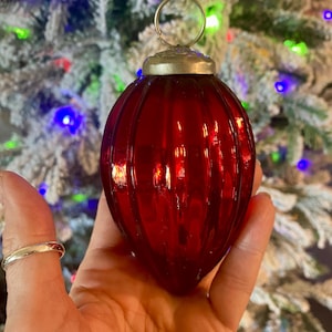 Może przedstawiać: Żywa czerwona ozdoba choinkowa z żebrowanym wzorem. Szklana ozdoba ma srebrną metalową nasadkę i pętlę do zawieszania. Tło przedstawia oszronioną choinkę z kolorowymi światłami.