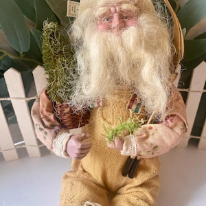 May include: A handcrafted figurine of an elderly man with a long white beard, wearing a straw hat and yellow dungarees. He holds a small green tree in a basket and a bundle of twigs. The figure has a detailed face and is dressed in rustic clothing.