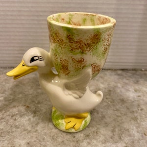 May include: A ceramic egg cup in the shape of a white duck with a yellow beak and feet. The egg cup is a light beige color with green and brown speckled patterns. The duck is standing on a green base.