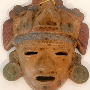May include: Handmade terracotta mask with a weathered appearance. The mask features a stylized face with open eyes and mouth, adorned with decorative elements in red, blue, and green. A yellow string is attached at the top for hanging. The mask is a unique piece of art.