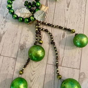 May include: A festive Christmas ornament featuring a green beaded ring with silver leaf accents and dangling green glass ball ornaments. The ornament has a rustic, vintage aesthetic, perfect for holiday decor.