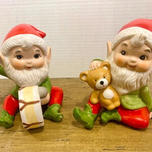 Könnte beinhalten: Zwei Keramik-Weihnachtselfen-Figuren. Einer spielt eine Trommel mit einem beige-braunen Design, der andere hält einen kleinen Teddybären. Beide Elfen tragen rote Hüte, grüne Jacken und rote Hosen.