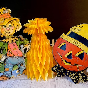 May include: A festive Halloween display featuring a scarecrow, a honeycomb decoration, and a jack-o'-lantern. The scarecrow wears a straw hat and green coat. The jack-o'-lantern has a black hat and bow tie. The honeycomb is yellow.