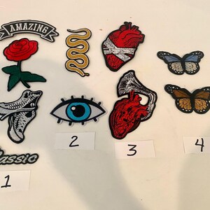 May include: A collection of embroidered patches featuring a red rose with the text AMAZING, a yellow snake, a red anatomical heart with white bandages, two butterflies, a high-top sneaker, a blue eye, and a red heart with a gramophone horn.