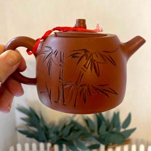 Puede incluir: Pequeña tetera de cerámica marrón con tapa y boquilla. La tetera presenta un diseño de bambú grabado y una cinta roja atada a la tapa. El asa es curva. La tetera se sostiene en una mano.