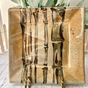 以下が含まれることがあります： 茶色と緑の色合いの竹模様が施された正方形のセラミックプレート。プレートはまだらで質感のある表面を持ち、金色の金属製のスタンドで支えられています。このプレートは装飾品です。