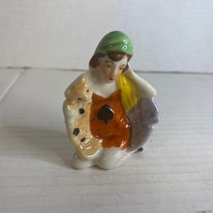 Könnte beinhalten: Kleine, vintage Porzellanfigur einer sitzenden Frau. Sie trägt ein grünes Kopftuch, ein orangefarbenes Kleid mit schwarzem Pik-Design und einen gemusterten Schal. Die Figur hat ein zartes, handbemaltes Aussehen.