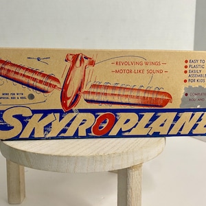 以下が含まれることがあります： SKYROPLANEというおもちゃの長方形の段ボール箱。箱はベージュの背景に青と赤の文字とグラフィックが特徴です。回転する翼を持つ赤いおもちゃの飛行機、ロッド、リールのイラストが描かれています。テキストには「REVOLVING WINGS」、「MOTOR-LIKE SOUND」、「EASY TO FLY」、「PLASTIC」、「EASILY ASSEMBLED」、「FOR KIDS 6 TO 60」、「COMPLETE WITH ROD AND REEL」が含まれています。