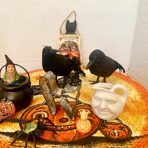 May include: A collection of Halloween decor including two black feathered crows, a black plastic cauldron with a small gnome figure, a metal chair, a white ceramic face pitcher, a glittery orange pumpkin, a black spider, and a cat sign.