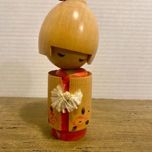 Pode incluir: Uma boneca Kokeshi de madeira com cabeça e corpo castanho claro. A boneca tem olhos fechados e um ponto vermelho no topo da cabeça. Ela usa um quimono vermelho com uma jaqueta amarela adornada com desenhos florais e um laço branco.