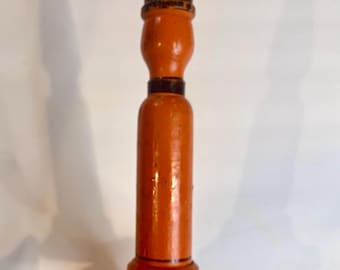 May include: Orange wooden candlestick with black accents. The candlestick has a flared base, a cylindrical body, and a tapered top. The paint shows some wear, giving it a vintage appearance. The candlestick is held in a hand.