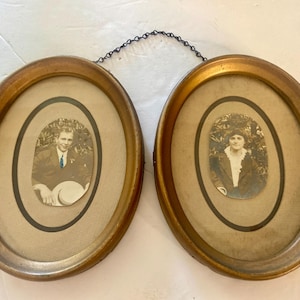 May include: Two oval gold-toned frames connected by a thin black metal chain. Each frame contains a beige mat with a black inner border and a vintage black and white portrait photograph of a person. The left portrait shows a person in a suit.