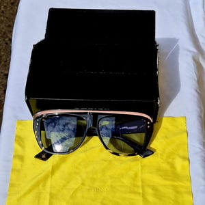 May include: Oversized sunglasses with a tortoiseshell frame, dark tinted lenses, and a pale pink top bar. The sunglasses are positioned in front of a black rectangular box and rest on a bright yellow microfiber cleaning cloth with the Dior logo.