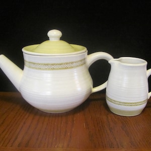 May include: A white teapot and creamer set with a green and brown geometric pattern. The teapot has a lid and a handle. The creamer has a handle.