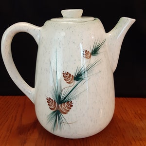 May include: White ceramic teapot with a speckled glaze and a pine cone design. The teapot has a brown handle and a lid.