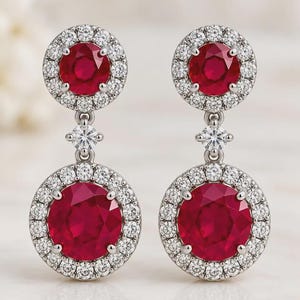 May include: A pair of drop earrings featuring two circular red gemstones, each surrounded by a halo of small white diamonds. A single small white diamond connects the upper and lower sections of each earring, set in polished silver metal.