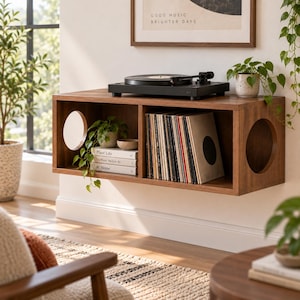 Peut inclure: Une unité de rangement murale en bois avec deux compartiments rectangulaires et des découpes latérales circulaires. Le dessus supporte une platine vinyle noire, tandis que les étagères intérieures contiennent des disques vinyles et trois livres intitulés Plant Life, The Music We Live By et At Home.