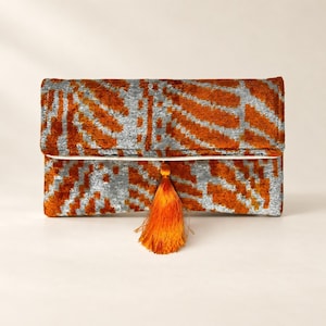May include: A rectangular clutch bag featuring a textured velvet fabric with a repeating abstract pattern in burnt orange and silver gray. The bag has a fold-over flap design and is accented with a single, vibrant orange tassel hanging from the center.