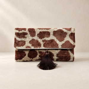 Puede incluir: Un bolso de mano rectangular con un tejido texturizado similar al terciopelo y un estampado animal inspirado en la jirafa en tonos marrón y crema. El bolso tiene un diseño de solapa plegable y está decorado con una borla de color marrón oscuro.