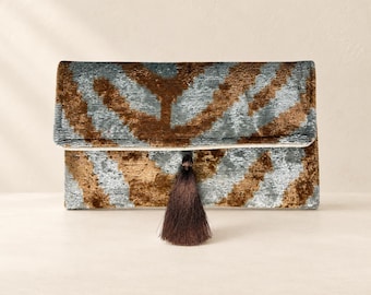 Puede incluir: Un bolso de mano rectangular con tejido de terciopelo texturizado y un patrón abstracto repetitivo en tonos plateado metálico y marrón oscuro. El bolso tiene un diseño de solapa plegable y está decorado con una borla decorativa marrón oscuro.