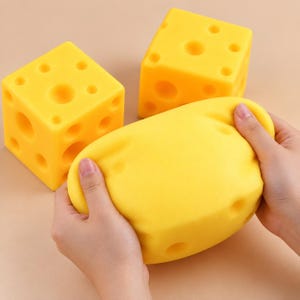 May include: Two bright yellow foam cubes shaped like Swiss cheese with various circular indentations and one squishy, flexible yellow foam piece being compressed by hands, demonstrating the soft and pliable texture of the material.